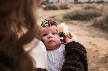 A woman holding a baby with a wreath of flowers on its head.