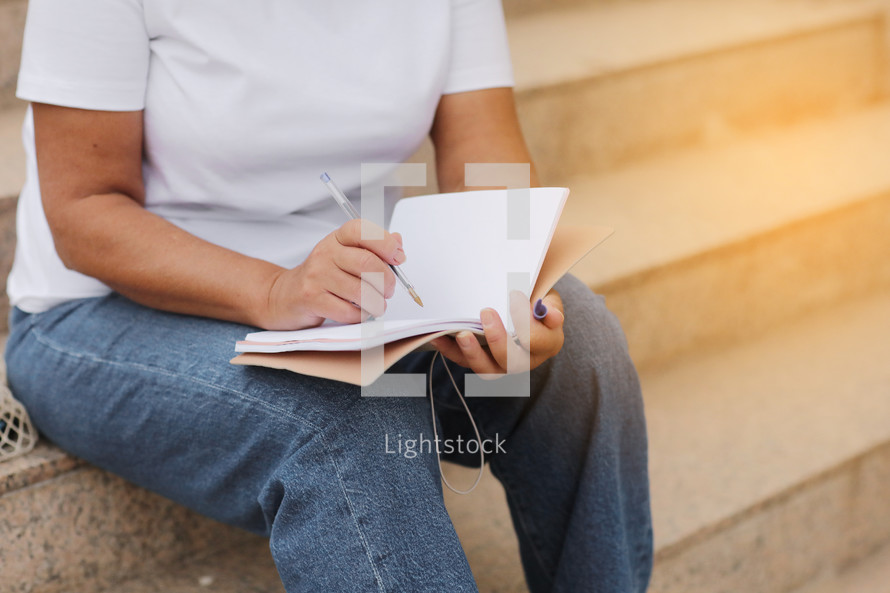 cropped photo woman writes and makes note in a notebook sitting on stairs outdoors. female works remotely,higher education for adults, freelancer. College study programs, academic educational courses
