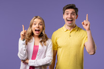 Happy stylish couple having idea eureka moment, pointing fingers up on violet background. Clever creative woman and man showing answer gesture, memory concept. High quality