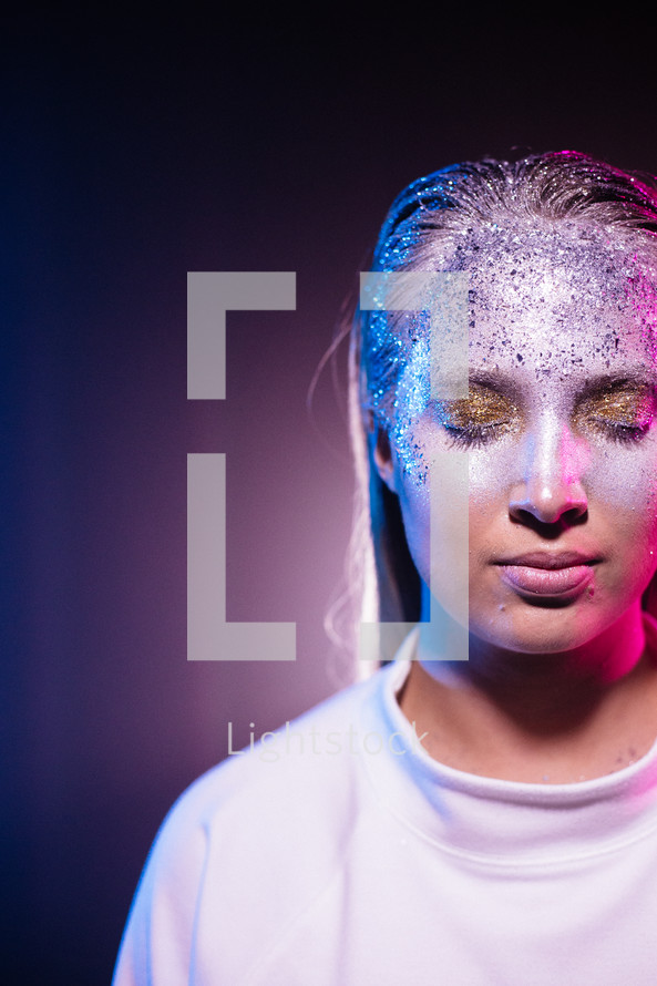 Woman standing in bright light with gold and silver makeup and glitter in her face. Colors in blue and pink wearing white clothing symbolizing an angel in modern shape