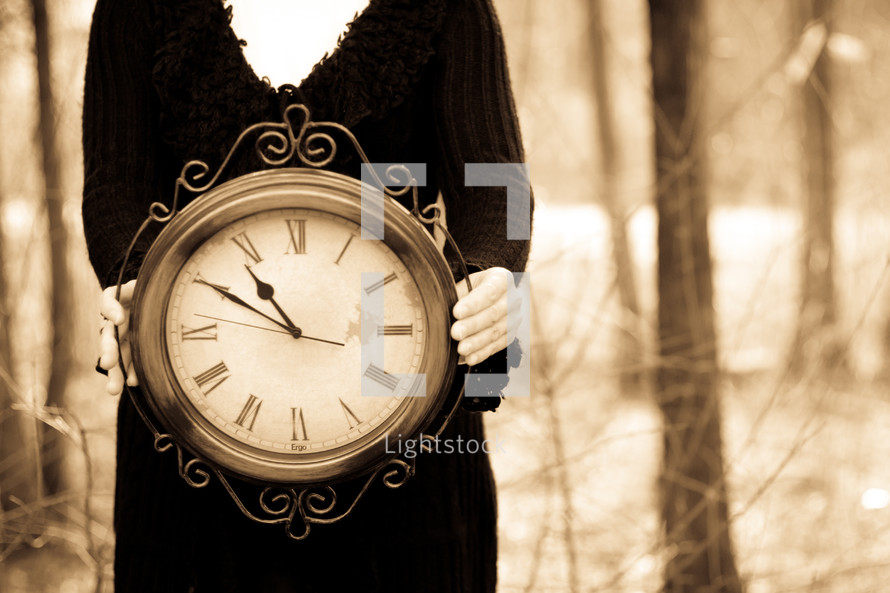 Woman in a black dress holding a large, ornate clock in a wooded area