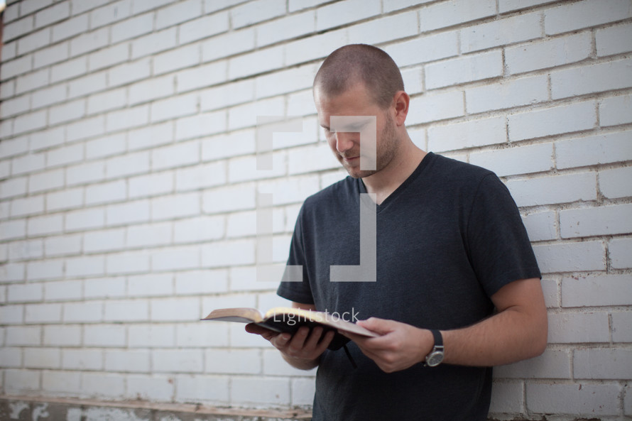 a young man reading a Bible standing in front of a white wall 