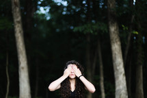 a woman standing in a forest covering her eyes 
