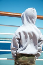 boy child in a hoodie looking through the rails of a ship out at the ocean