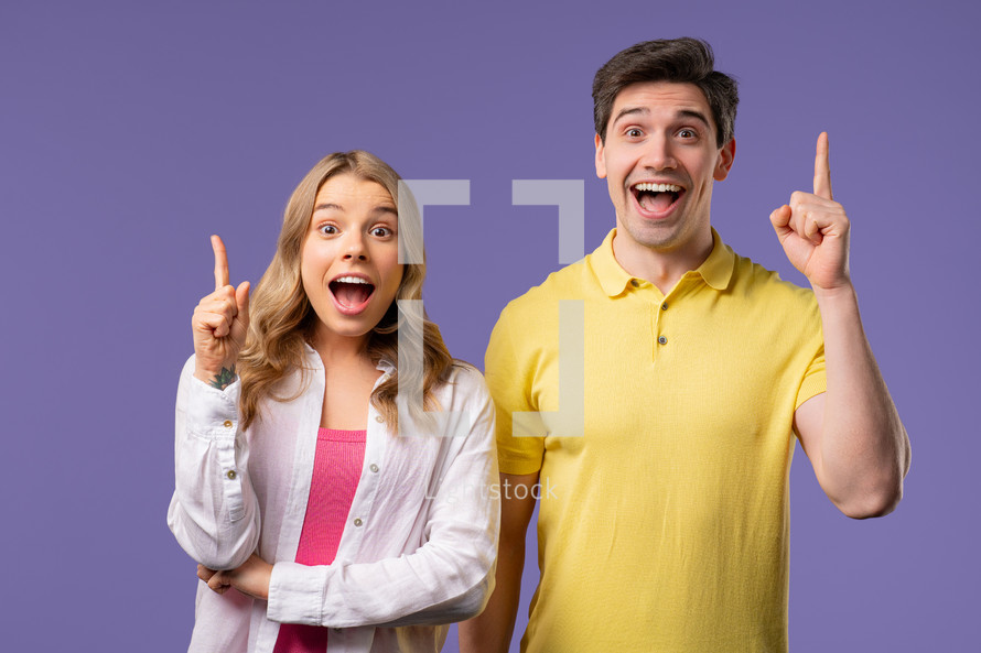 Happy stylish couple having idea eureka moment, pointing fingers up on violet background. Clever creative woman and man showing answer gesture, memory concept. High quality