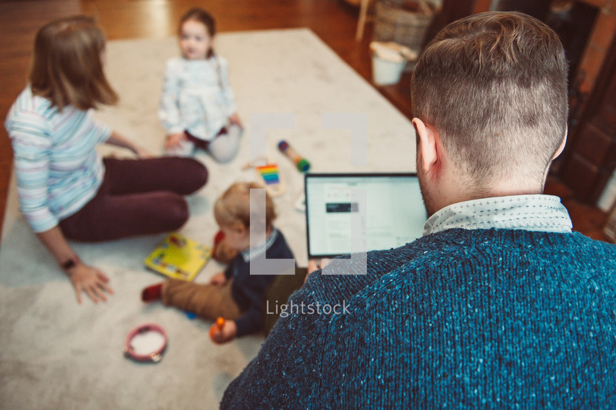a father looking at a tablet screen while his family plays on the floor under him