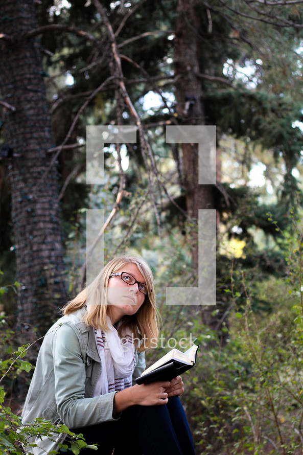 A young woman reads her Bible and thinks in a forest.