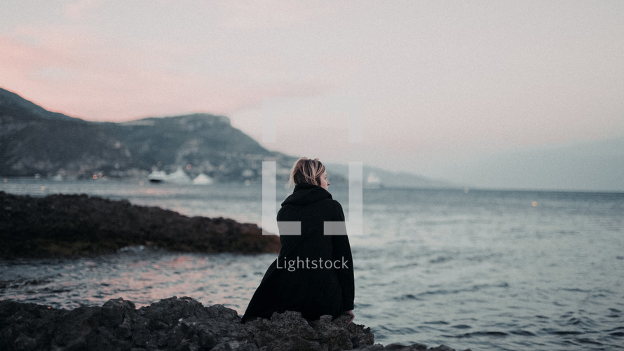 a woman sitting on a rocky shore in a coat 