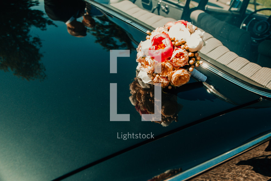 a bouquet of flowers on the hood of a car and bride and groom reflection 