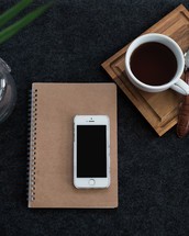 A cell phone on a spiral notebook next to a cup of coffee and wristwatch on a wooden coaster.