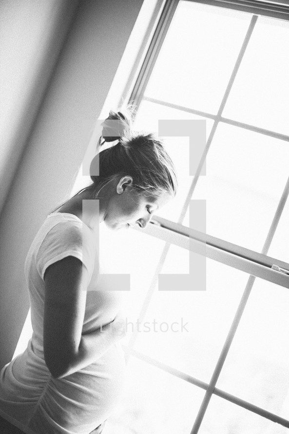 Pregnant woman standing in front of a window looking at her belly.