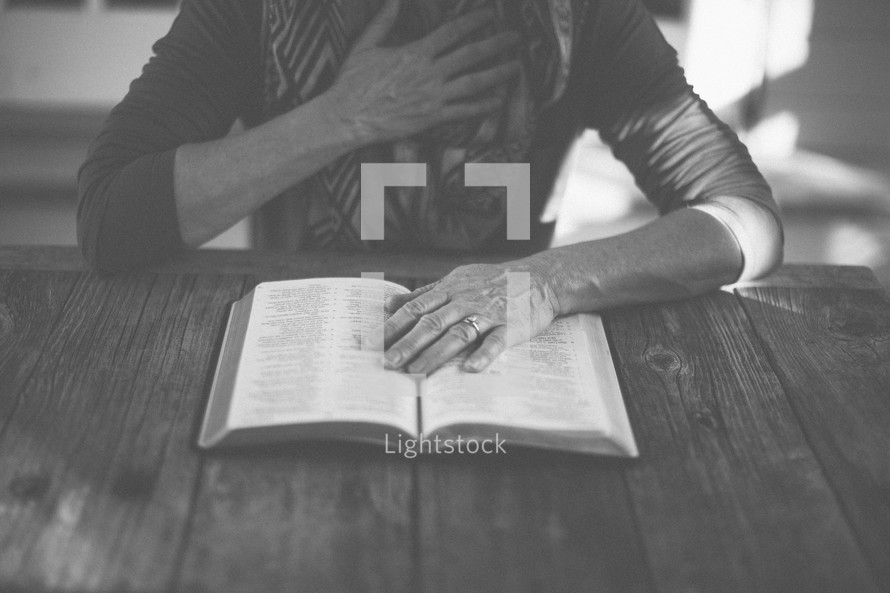 A woman reading the Bible at a table with her hand over her heart