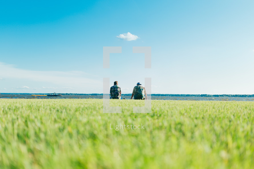 Two men sitting by a lake next to each other with a grassy foreground