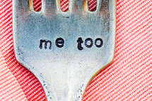the words me too engraved stamped into a dinner fork sitting on a pink linen