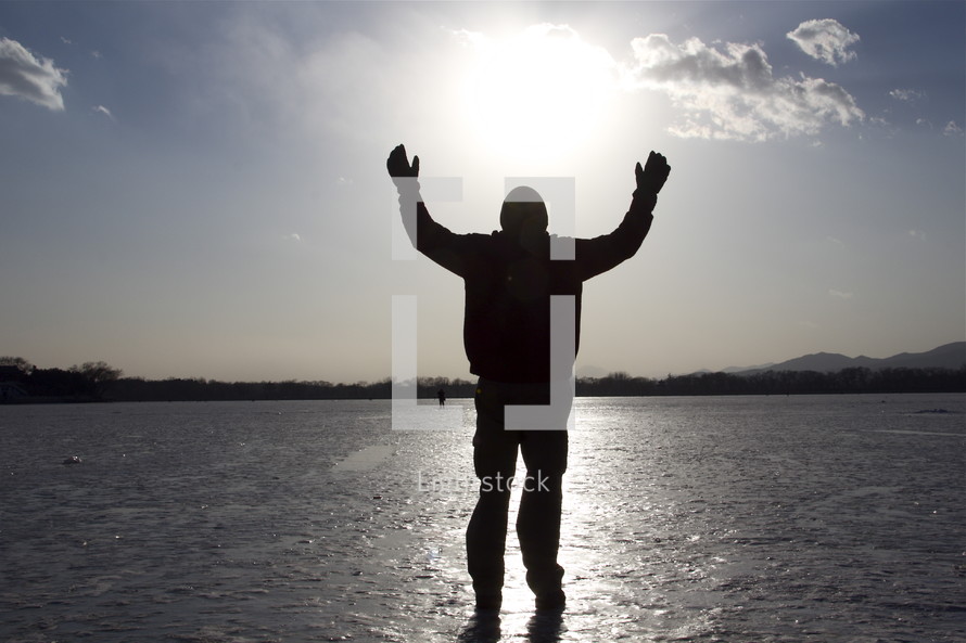 Silhouette of man with arms raised praising God while walking on ice over a frozen lake.