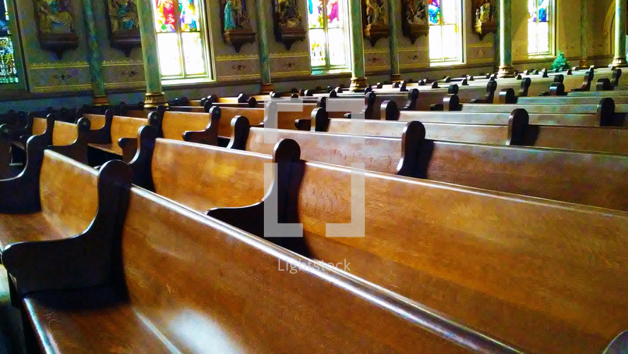 Rows of beautiful wooden church pews sit empty in a large church filled with stained glass windows and ornate architecture. 