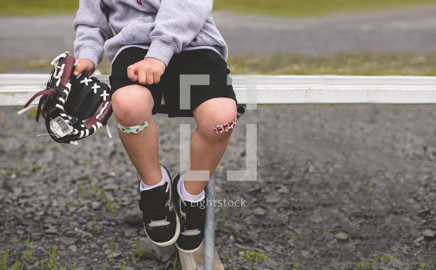 boy child sitting on a bench holding a baseball glove with band-aides on his knees