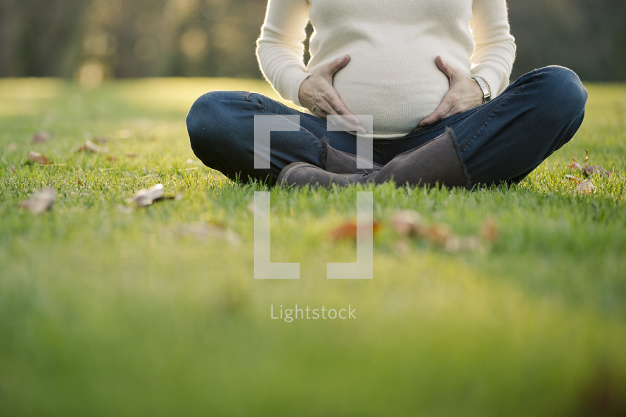 torso of a pregnant woman sitting in the grass with her hands on her belly
