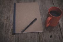 A pen and notebook next to an orange coffee mug