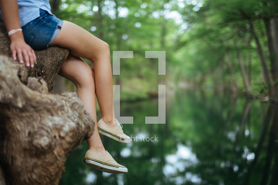 A girl in shorts dangles her legs from a tree over a river.