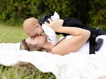mother kissing her baby boy while lying on a blanket in the grass