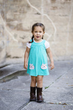 A little girl in a blue dress and boots standing in front of a stone wall.