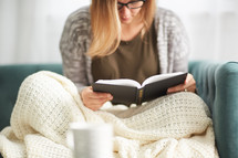 a woman reading a book on a couch with a blanket 