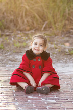 A young child in a red coat sitting on a brick path.