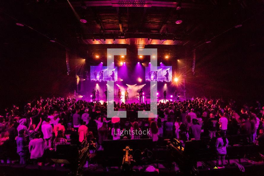 A large congregation in front of stage of musicians lit by colorful lights.