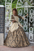 A woman dressed in colonial style dress, standing in front of a white, ornate gate, reading a book