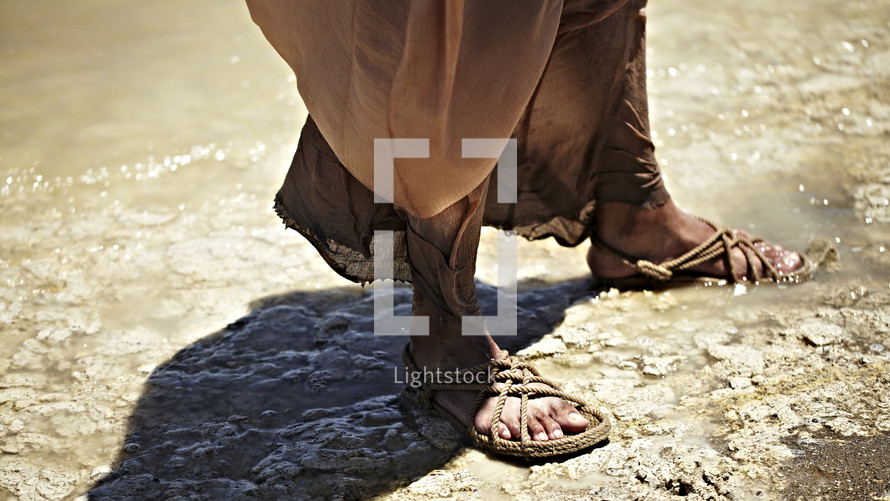 Jesus standing near the water with his feet in sandals.