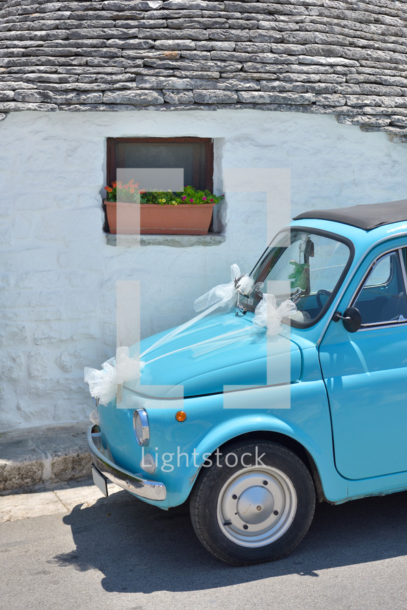 Old light blue tiny cute italian car decorated for marriage parcked outside an old trulli house from Alberobello, south Italy