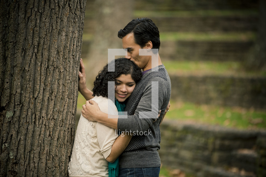 Man kissing woman on top of the head as they lean against a tree.