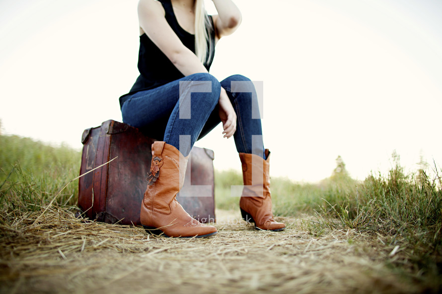 woman sitting on a suit case on a dirt road boots grass