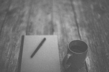 A pen and notebook next to a coffee mug on a wooden table