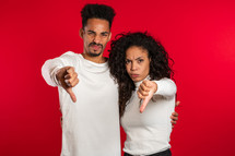 Young african couple standing on red studio background expressing discontent and showing thumb down gesture at camera. Portrait of man and woman with sign of dislike