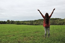 young woman standing in a field with arms raised 