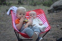 toddler brother and infant sister sitting together in a chair outdoors 