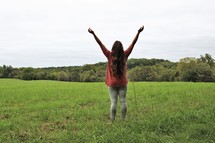 a young woman standing in a field with hands raised 