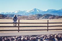 woman sitting on a fence looking out at a mountain 