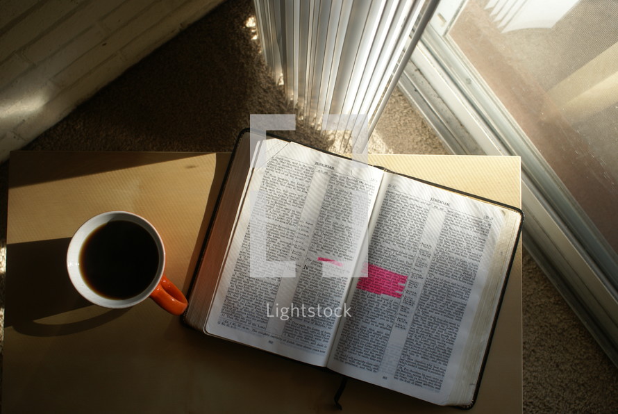 Aerial view of an open Bible on a table with a cup of coffee, by a window.