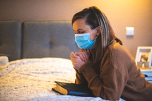 a woman praying bedside wearing a face mask 