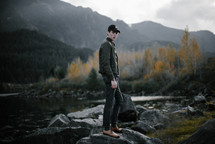 a young man in dress shoes standing on rocks by a lake 