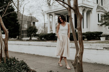 a woman in a white dress walking down a neighborhood sidewalk 