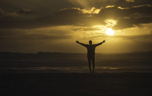 silhouette of a man with arms raised standing on a beach at sunset 