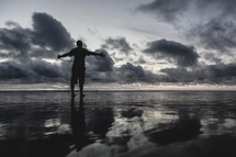 a man standing on a beach on wet sand with opened arms  at sunset 