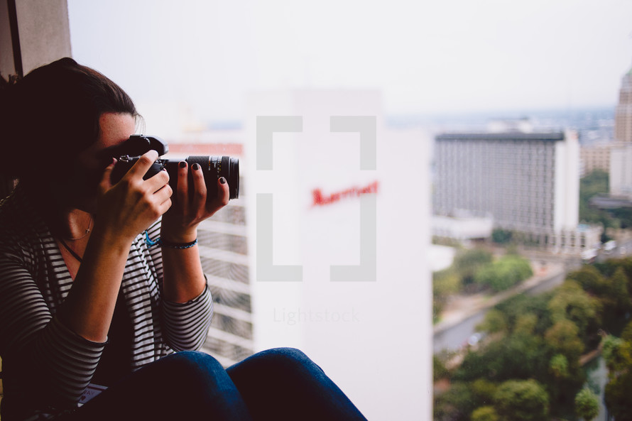 woman with a camera taking a picture while sitting in a window sill 