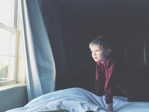 boy child on a bed looking out a window 