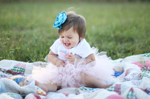 Smiling toddler girl in a tutu sitting on a quit in a field of grass.