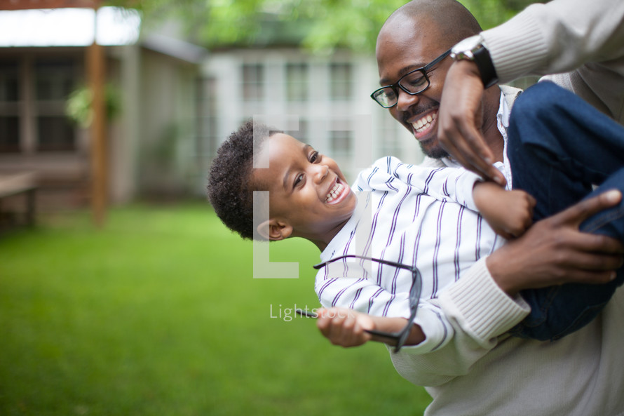 A father holding and playing with his young son in the backyard.